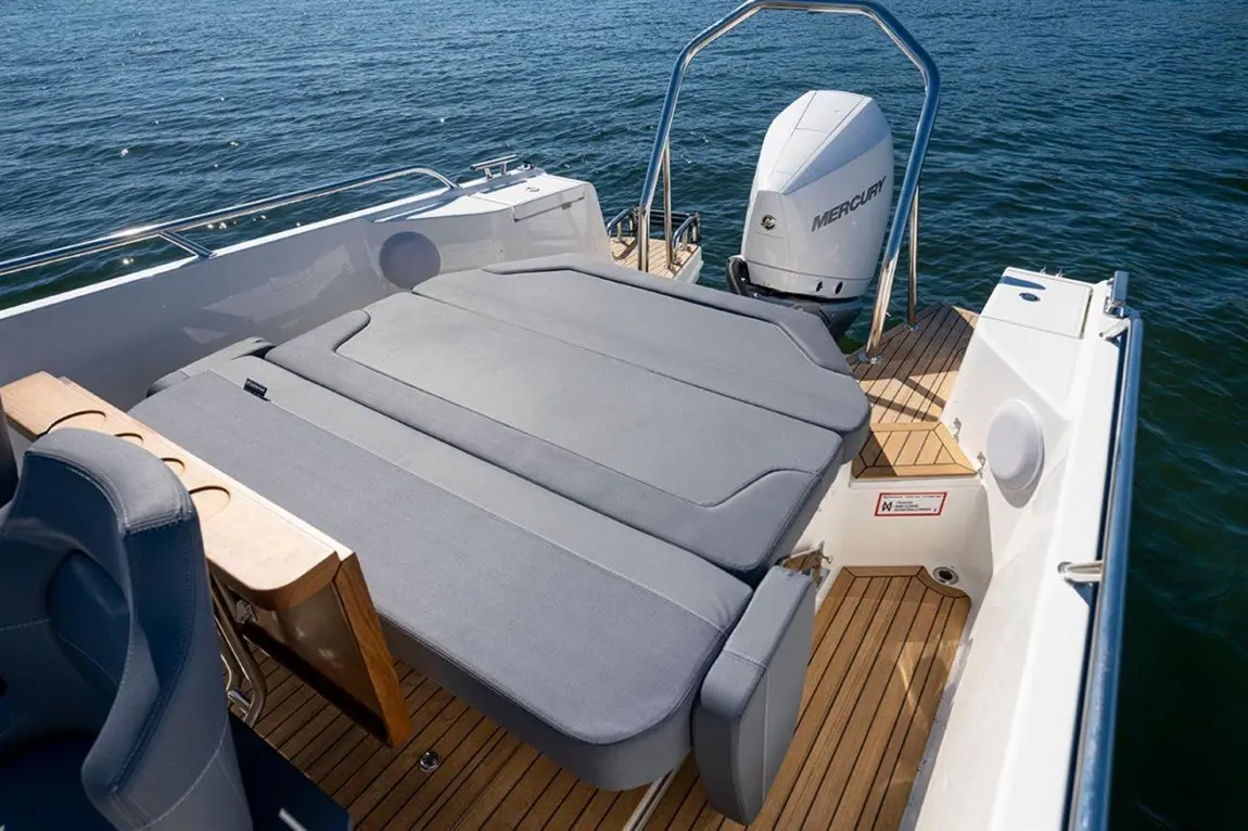 Interior of a boat featuring grey cushioned seating, a wooden table, and a Mercury outboard motor, with water in the background.